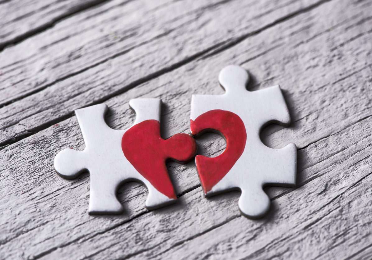 closeup of two separated pieces of a puzzle which together form a heart on a white rustic wooden surface, depicting the idea of rupture or cooperation